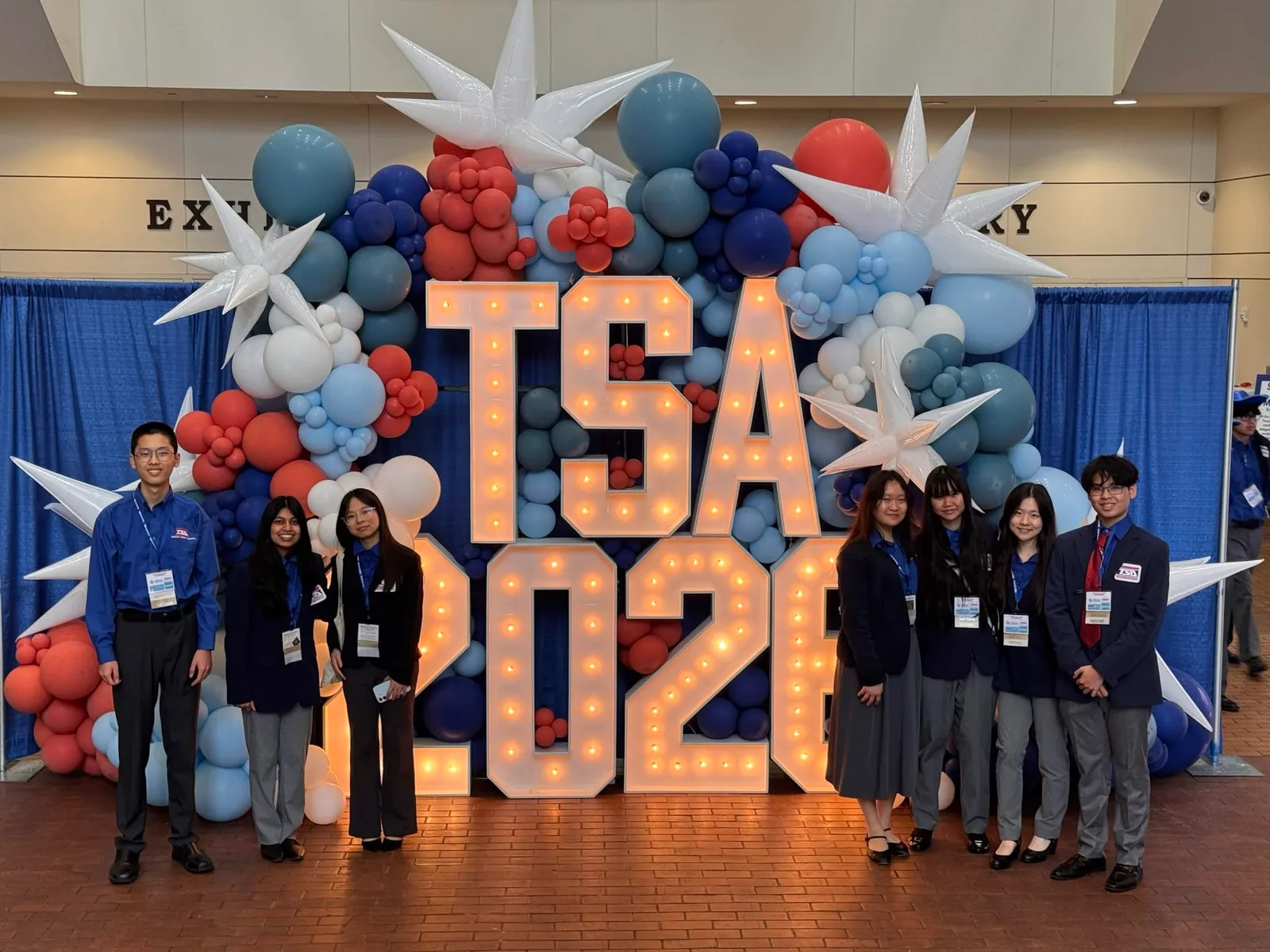 Members in conference dress standing on both sides of the giant light-up TSA 2026 marquee letters with red, white and blue balloon garland at the Fort Worth Convention Center